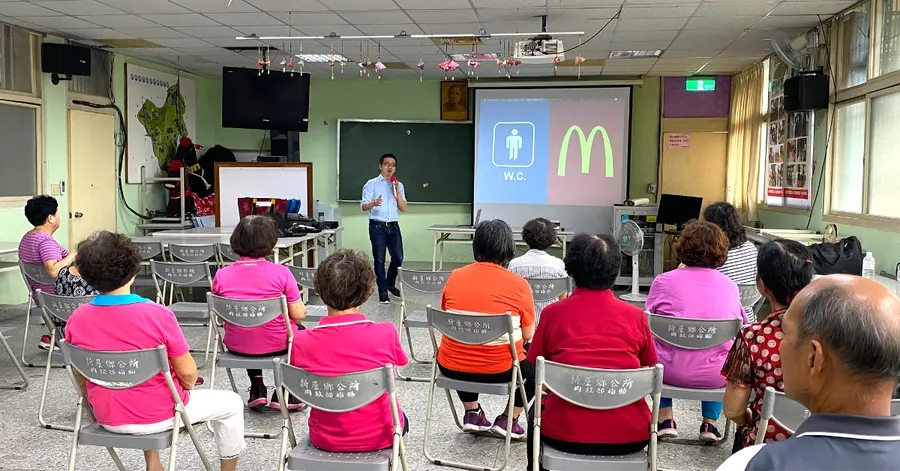 社區衛教的現場,長輩們坐在台下,投影幕上是廁所標誌與速食店招牌的對照。